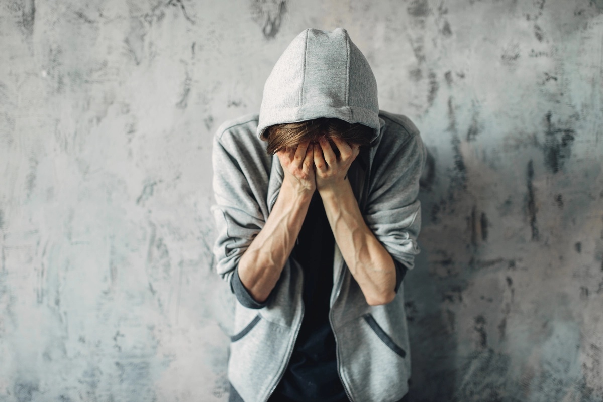 A young man dealing with cocaine addiction stands against a wall, holding his hands to his face.