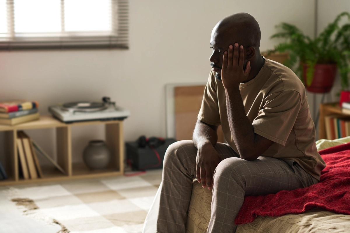 persistent-depressive-disorder A man sitting on the edge of his bed with his hand up to his face in a sad manner, possibly struggling with symptoms of persistent depressive disorder.