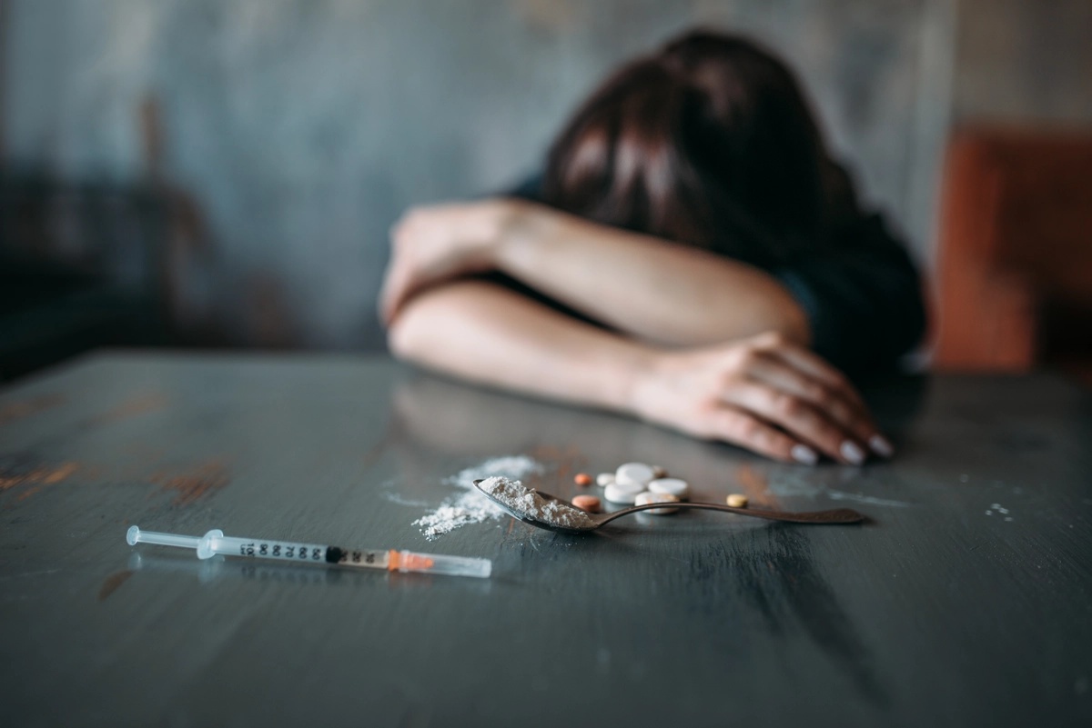 A person struggling with meth addiction lays her head on her arms at a table, with a needle and meth also on the table. A person struggling with meth addiction lays her head on her arms at a table, with a needle and meth also on the table.